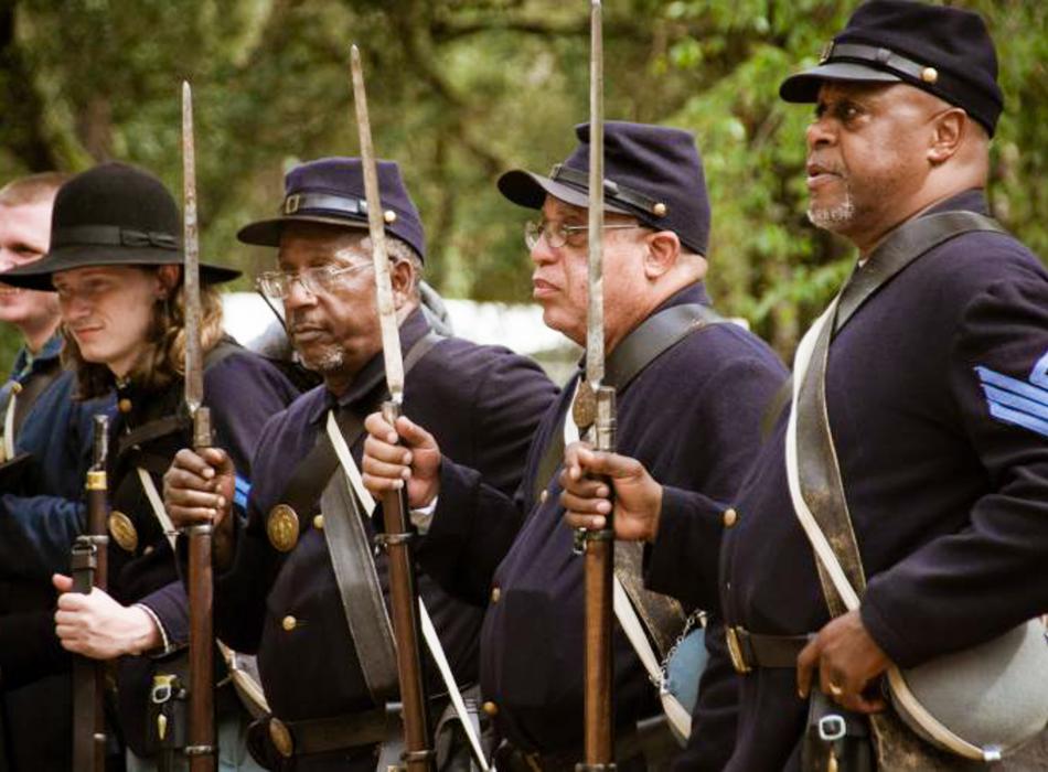 Black Union reenactors stand shoulder to shoulder, looking on with muskets in hand.